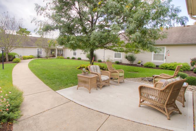 A serene outdoor patio with wicker chairs set on a concrete path surrounded by lush grass and trees.