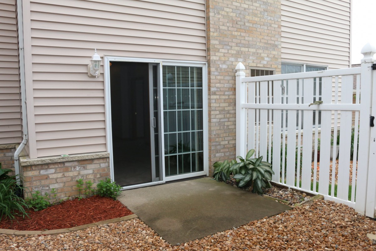 A small patio area with a sliding glass door, white fence, and some plants.