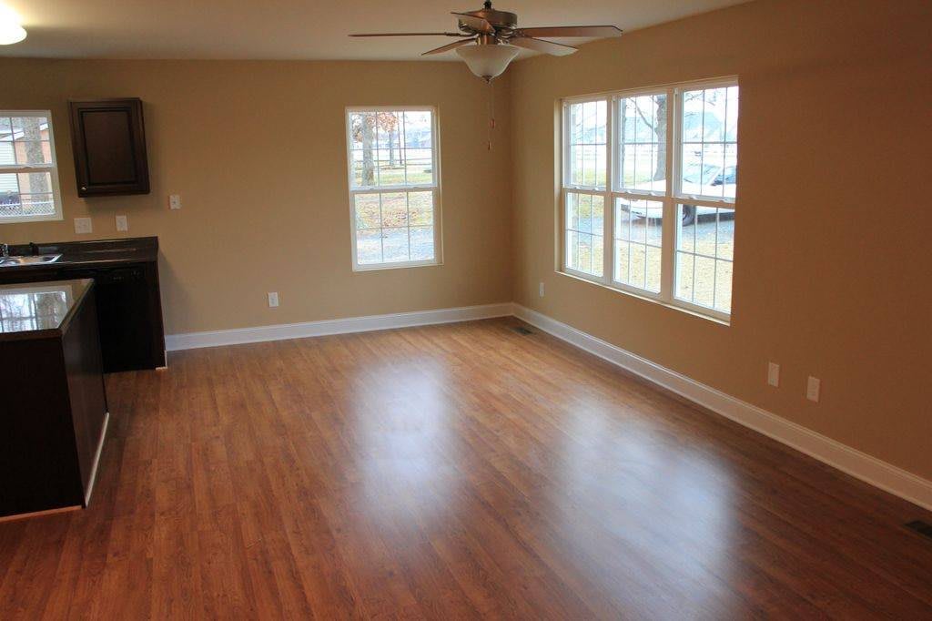 A spacious empty room with wooden flooring, large windows, and a ceiling fan.