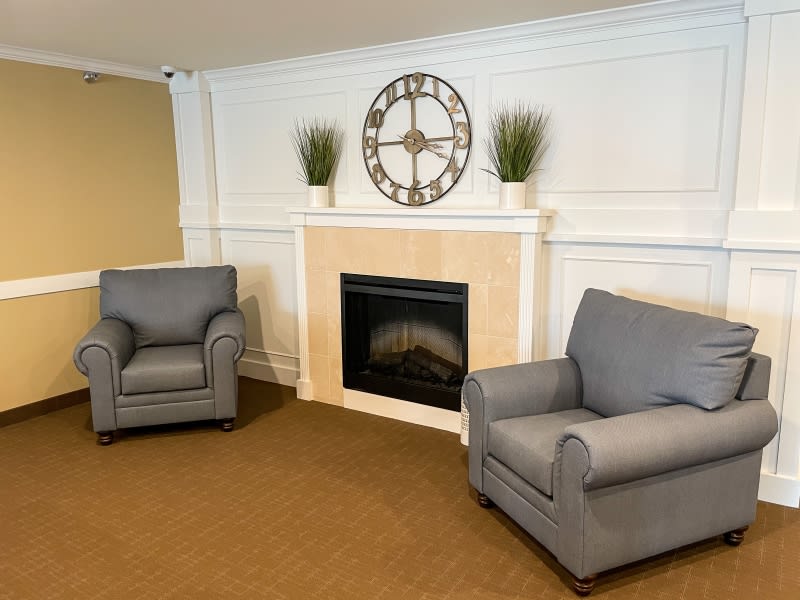A cozy living room with two gray armchairs facing a fireplace, adorned with decorative plants and a clock.