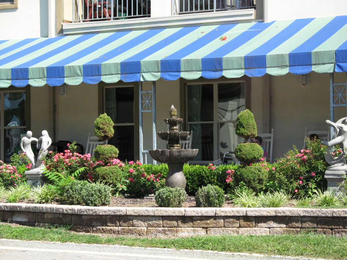 A charming garden with a striped blue and green awning, featuring a central fountain surrounded by topiary shrubs and colorful flowers.