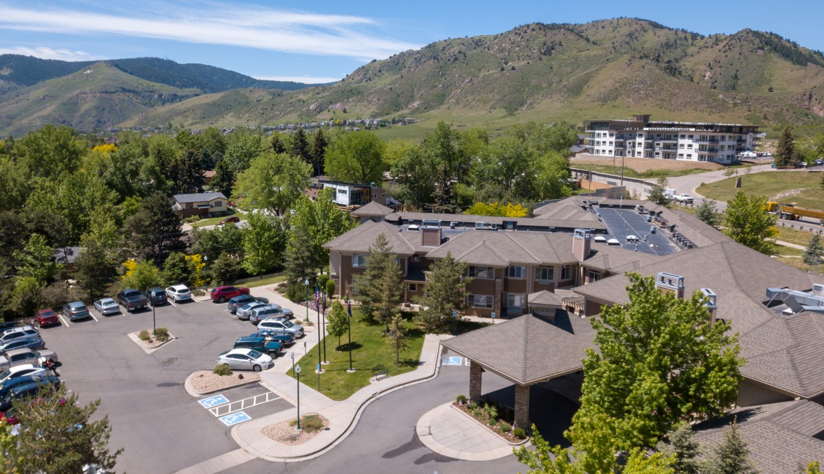 Aerial view of a building surrounded by a parking lot and lush greenery, set against a mountain backdrop on a sunny day.
