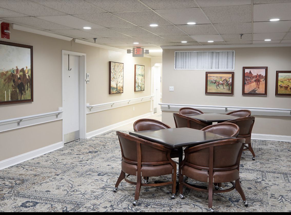 A quiet room with upholstered chairs and tables, decorated with paintings and carpeted flooring.