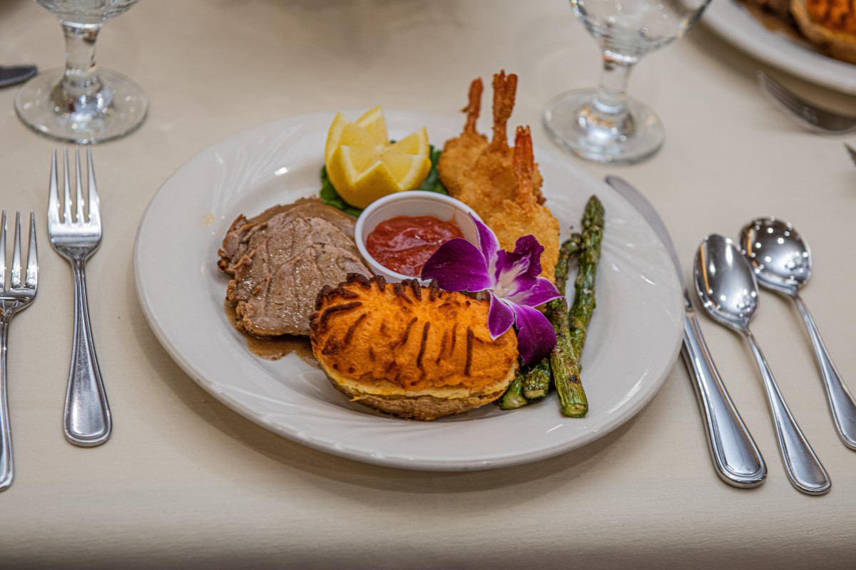 A gourmet surf and turf dish featuring steak, lobster tail, fried shrimp, and grilled asparagus, elegantly presented with a lemon wedge and sauce on the side.