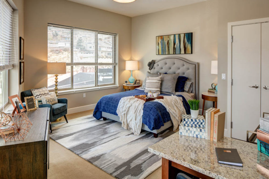A cozy bedroom featuring a tufted headboard, blue bedding, and warm lighting from bedside lamps. The room is completed with a large window and modern artwork on the wall.