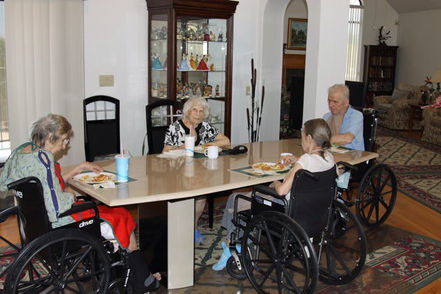 Elderly individuals sitting in wheelchairs around a dining table, enjoying a meal together indoors.