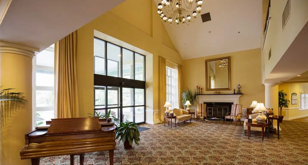 A spacious and elegantly designed living room featuring a piano, chandelier, fireplace, and large windows.