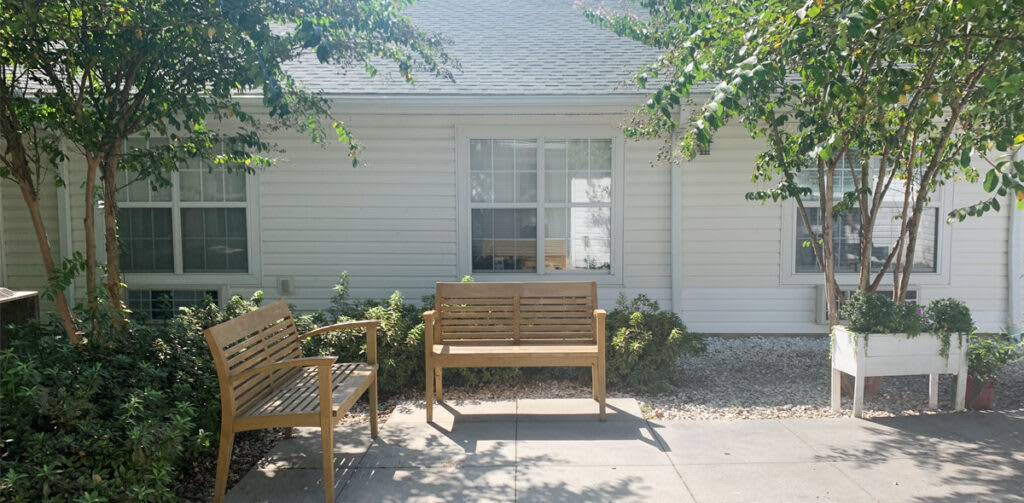 A sunny outdoor seating area in front of a house with wooden benches and trees providing shade.