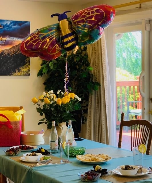 A festive indoor party setup with a colorful balloon decoration and a table set with food and flowers.