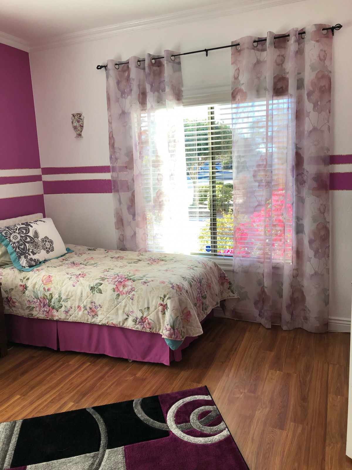 A cozy bedroom featuring a single bed with floral bedding and purple decor, complete with a window adorned with sheer curtains, allowing sunlight to illuminate the wooden flooring.