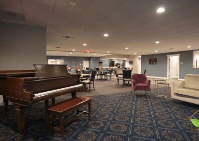 A spacious room with a piano and various seating arrangements, featuring a patterned carpet and warm lighting.