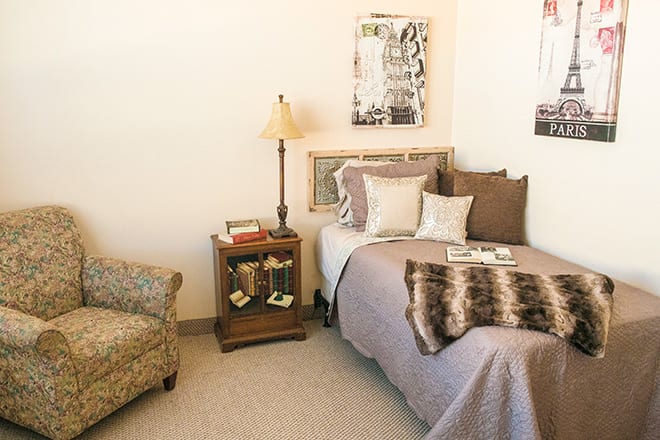 A cozy bedroom featuring a single bed with pillows and a blanket, an elegant nightstand with a lamp, and an armchair.
