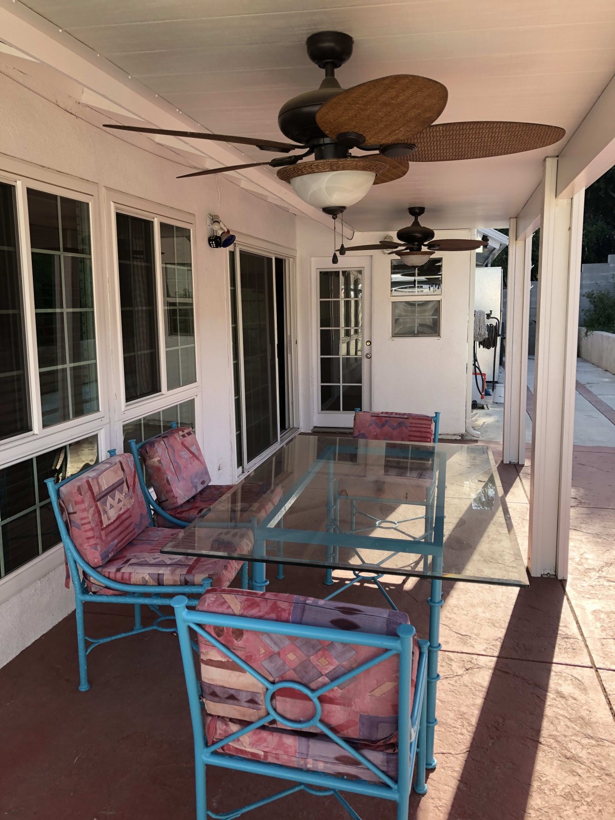 A cozy patio area with a glass table surrounded by cushioned chairs, under ceiling fans and near large windows.