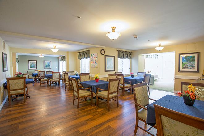 A spacious dining room with neatly arranged tables and chairs on a wooden floor, illuminated by ceiling lights and natural light from large windows.