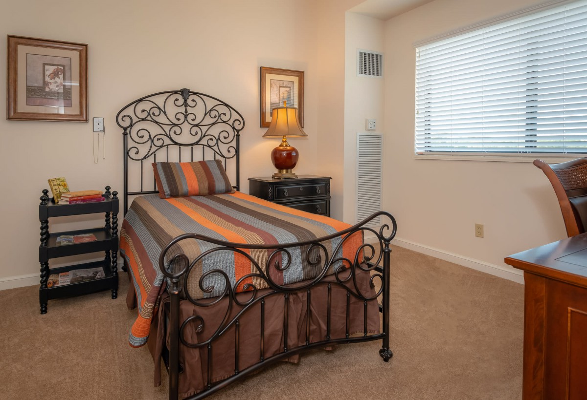 A cozy bedroom featuring a single bed with decorative metal frame, a nightstand with a lamp, and a window with blinds.