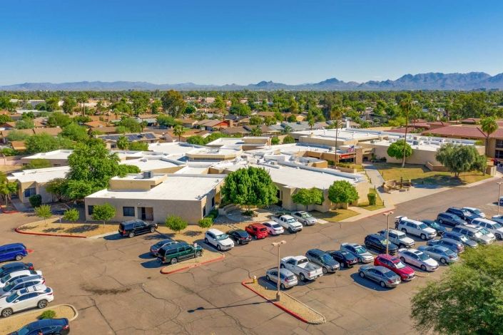 Aerial view of a suburban area with a parking lot and buildings surrounded by trees, set against a backdrop of mountains and clear blue sky.