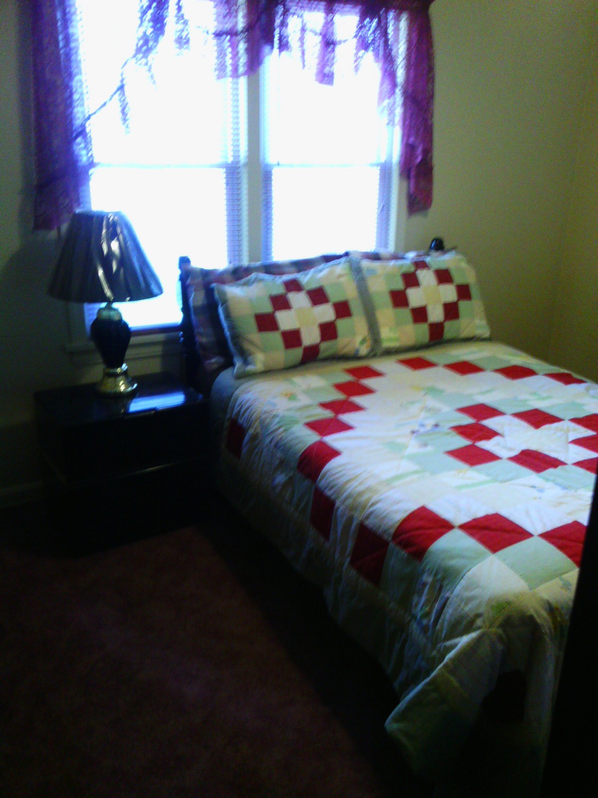 A cozy bedroom with a bed featuring a patterned quilt, pillows, and a bedside lamp on the nightstand next to a window with curtains.