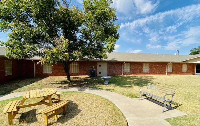 A brick house with a yard featuring a tree and picnic bench under a blue sky.
