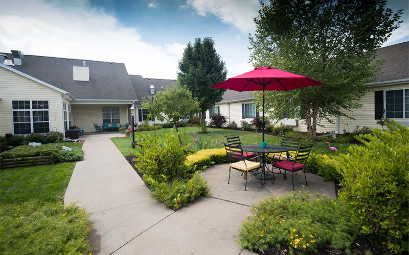 A scenic outdoor garden area with a patio, featuring a red umbrella and seating surrounded by lush greenery.