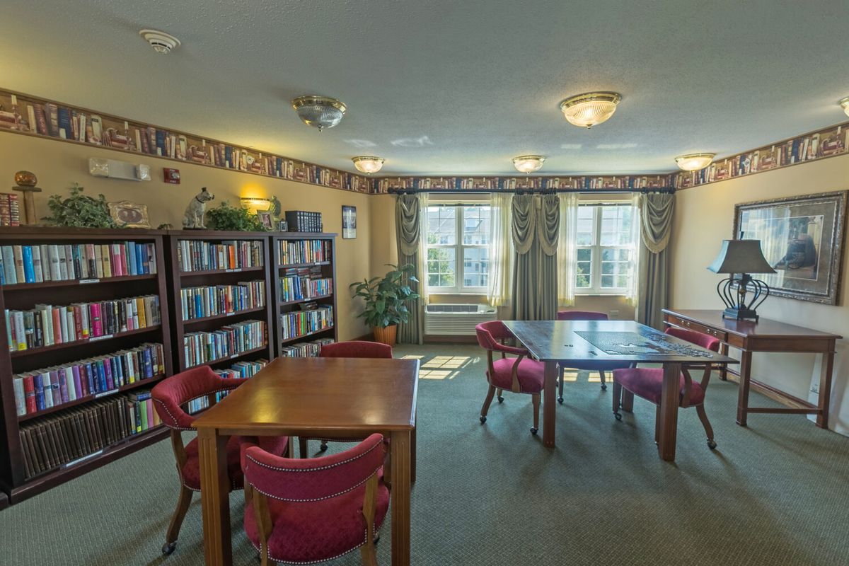 A cozy library reading room with bookshelves, tables, and chairs near large windows.
