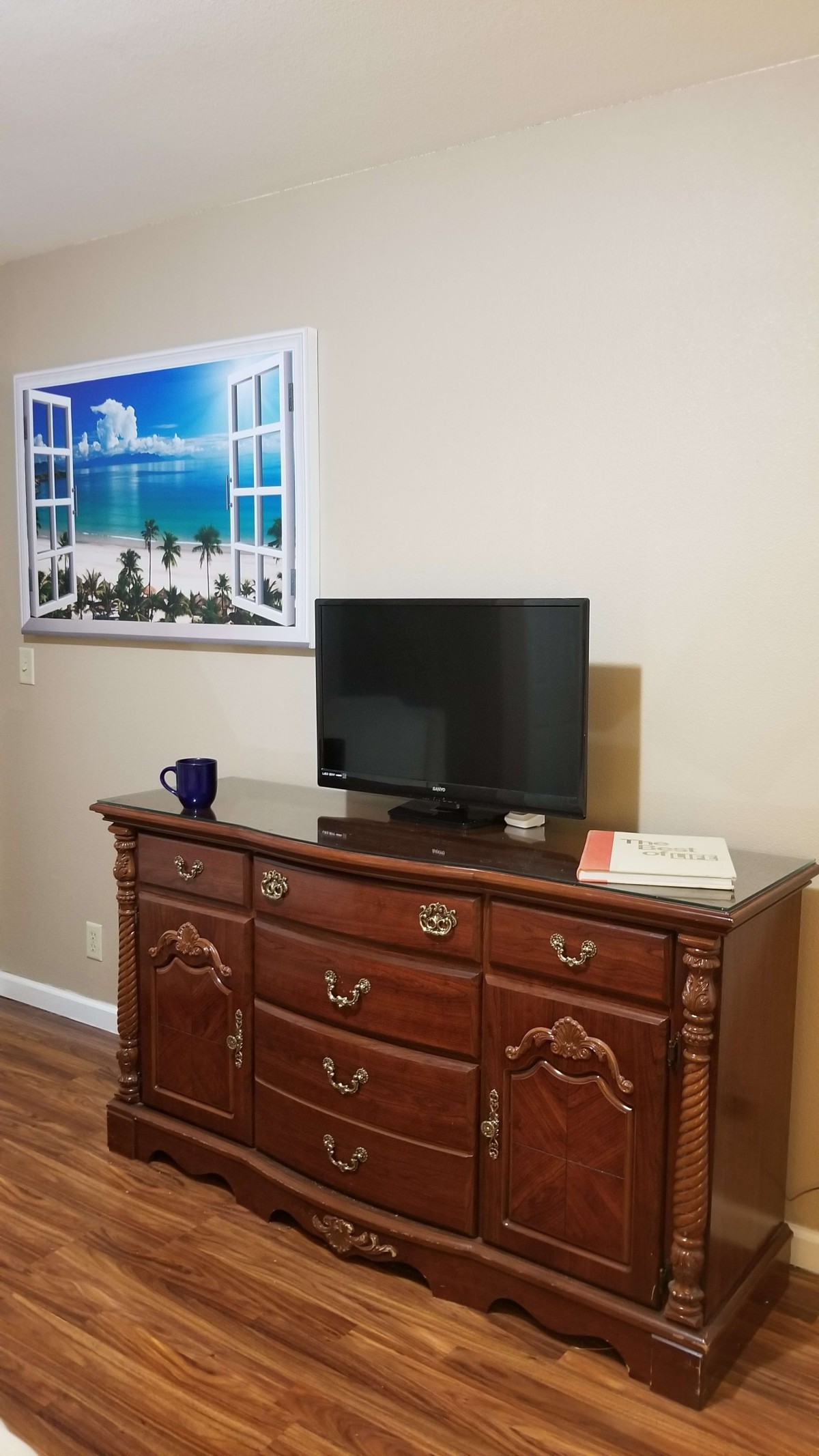 A room with a wooden dresser, a small television on top, and a framed artwork of a beach scene on the wall.