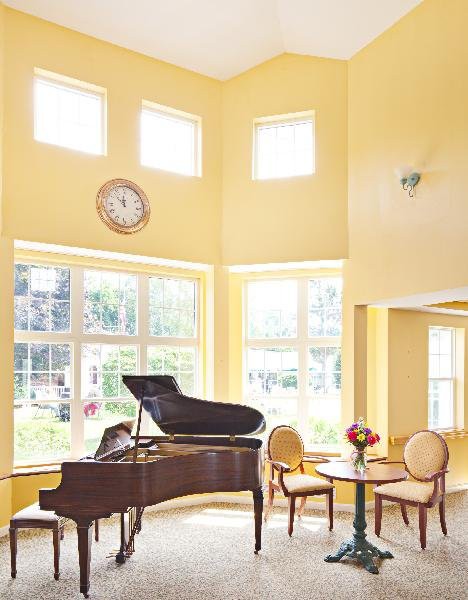 A bright living room with a grand piano, yellow walls, and abundant sunlight streaming through large windows.