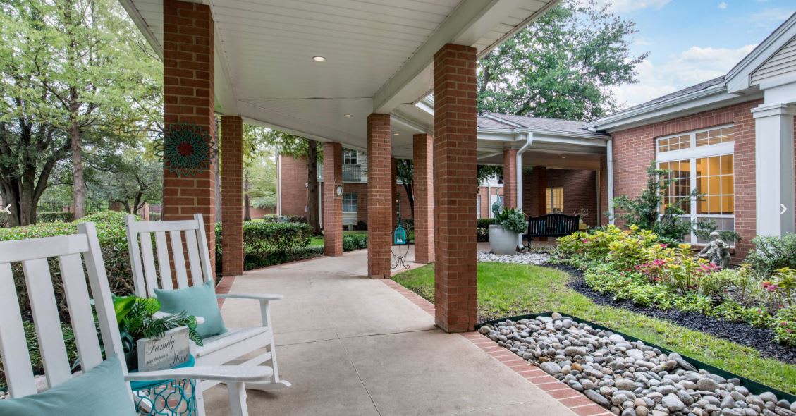 A peaceful outdoor patio with a covered walkway, surrounded by greenery and featuring brick columns and a rocking chair.