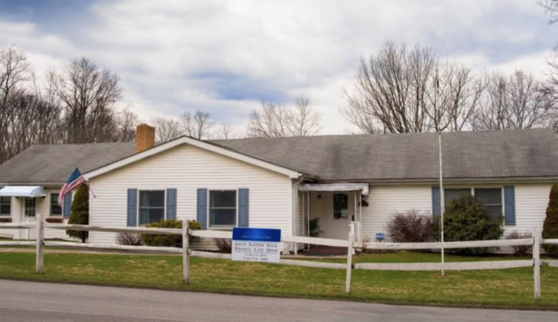 A single-story residential house with a white exterior and blue shutters, surrounded by a wooden fence, under a cloudy sky.