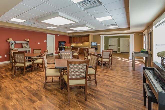 A spacious dining area with multiple tables and chairs, featuring a wooden floor and a kitchenette in the background.