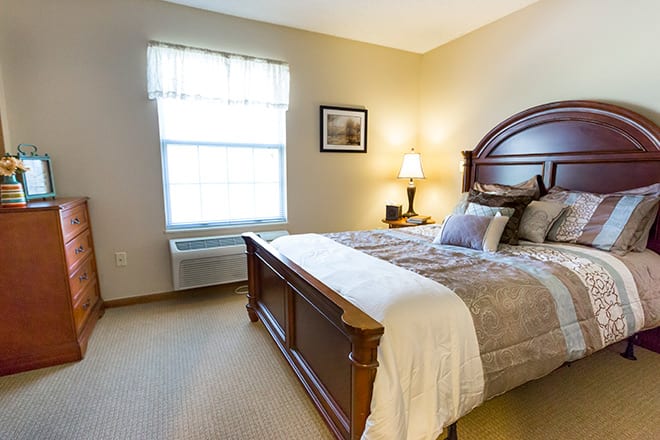 A cozy bedroom with a neatly made bed, bedside lamp, and natural light coming through the window.