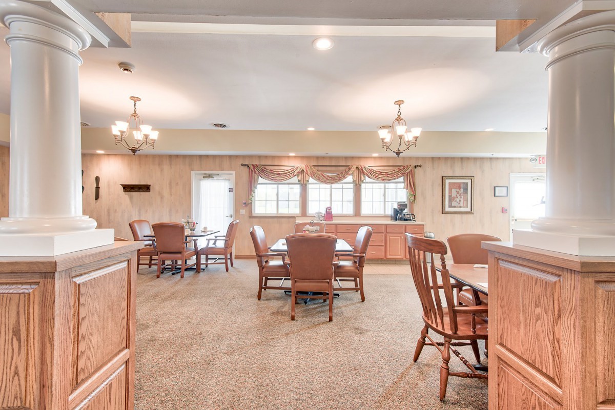 A spacious dining room with multiple tables and chairs, featuring chandeliers and natural lighting from large windows.