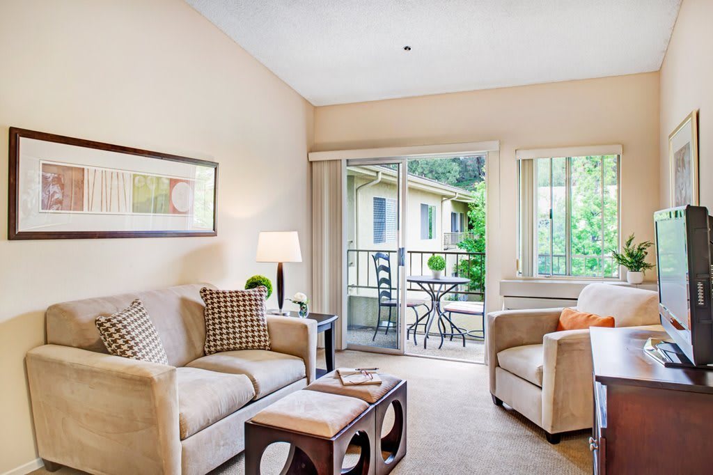 A cozy living room with neutral tones, featuring a sofa, armchair, and coffee table, opening to a balcony with outdoor seating.