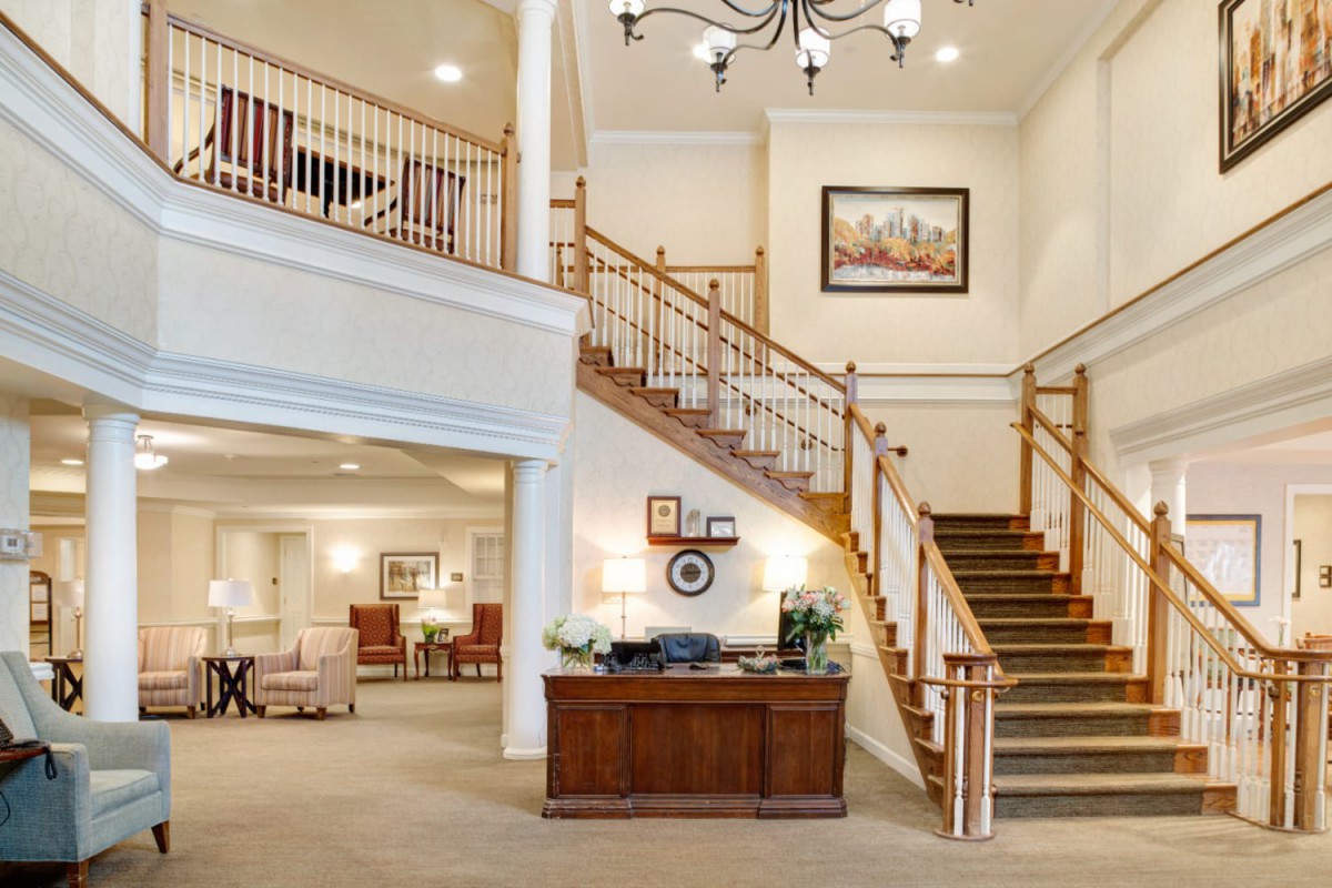 A spacious lobby featuring a wooden staircase and reception desk, adorned with a chandelier and artwork.
