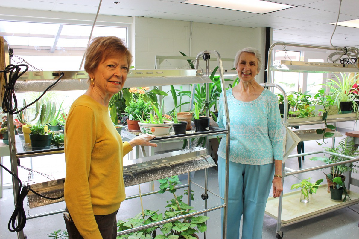 Two women smiling in a room filled with indoor plants and gardening equipment.