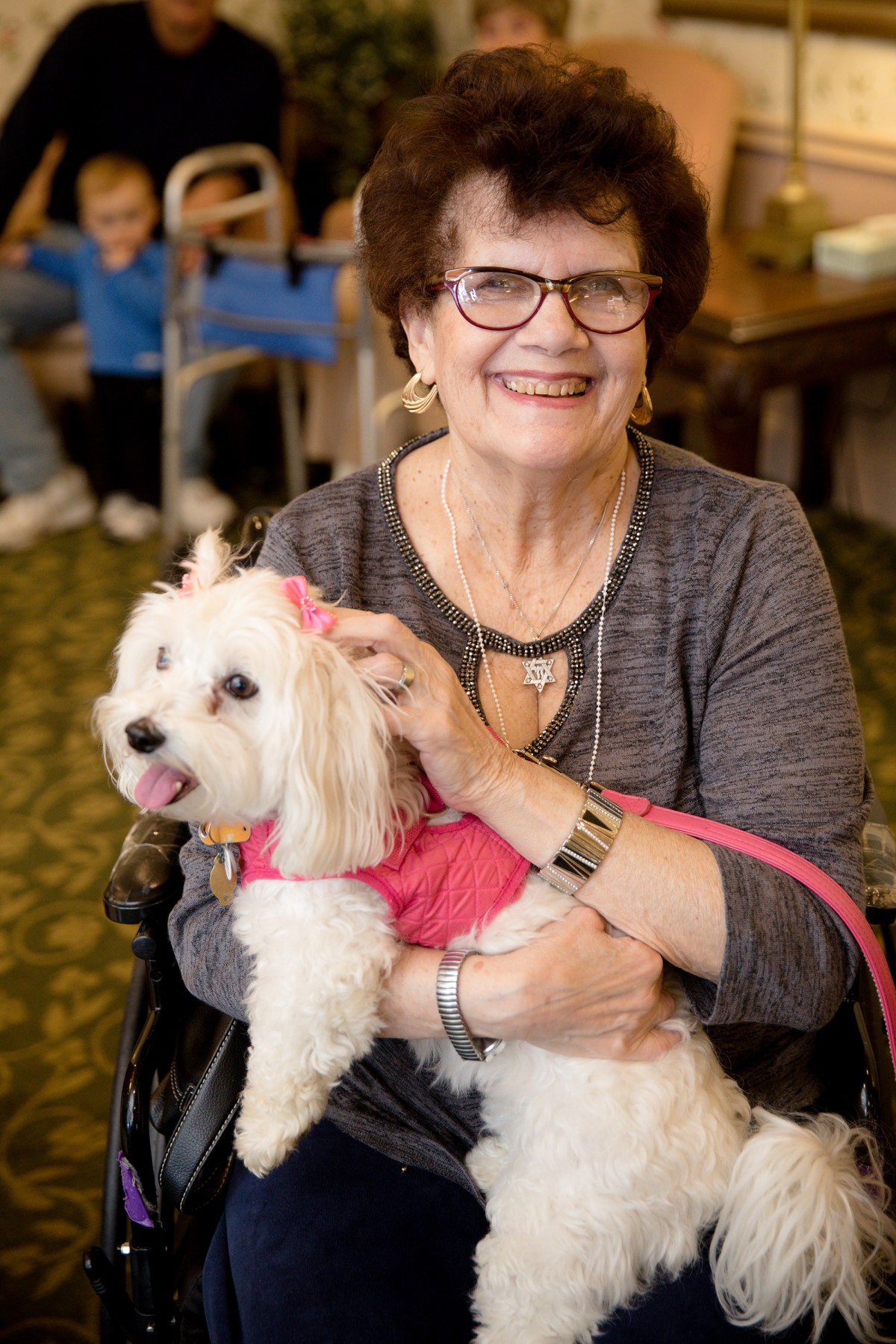 A smiling woman holds a small white dog wearing a pink outfit indoors.