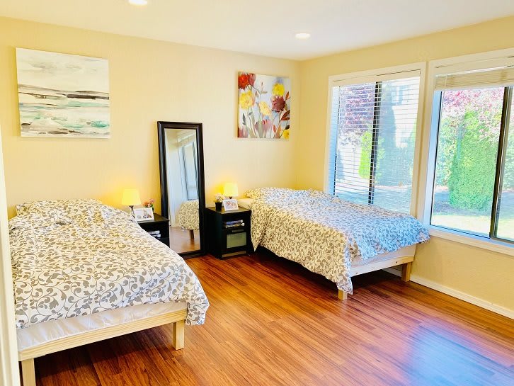 A bright bedroom with two beds and wooden flooring, featuring large windows and modern decor.