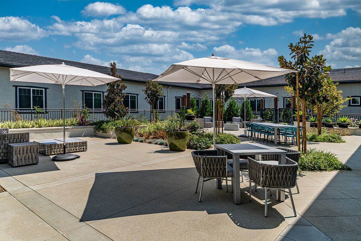 A sunny outdoor patio area with tables, chairs, and umbrellas surrounded by landscaping.