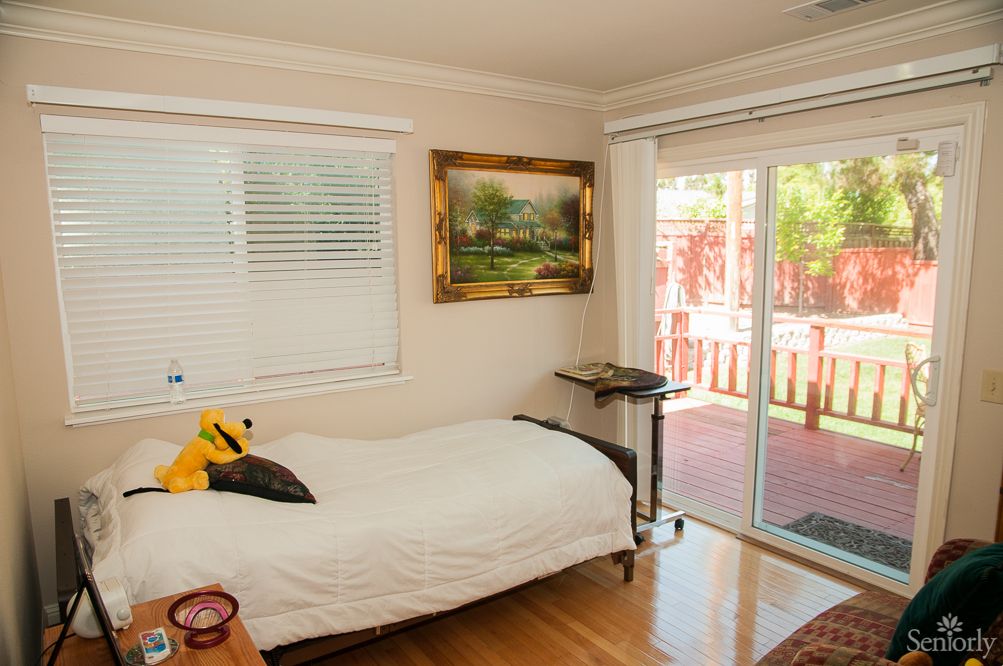 A cozy bedroom with a bed, painting on the wall, and sunlight streaming through a sliding glass door.