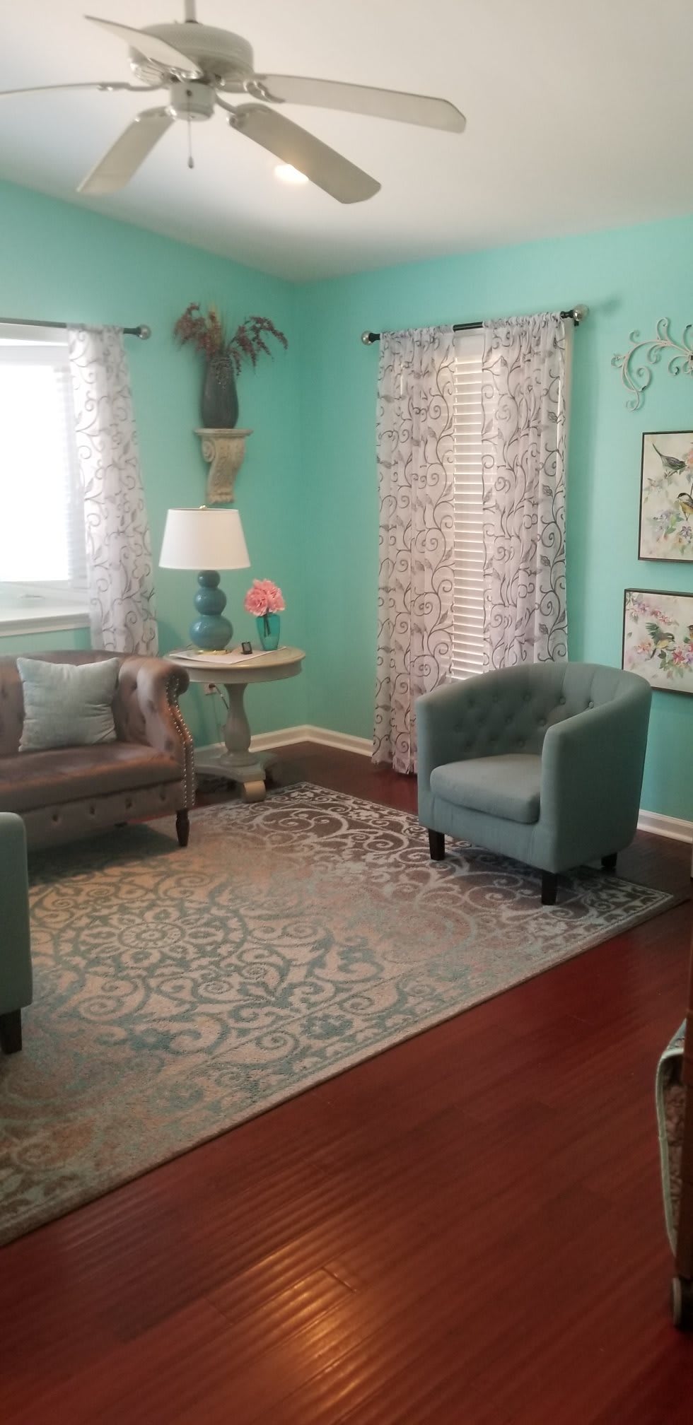A cozy living room with teal walls, featuring a comfortable armchair and sofa, accented by a patterned rug and decorative elements.