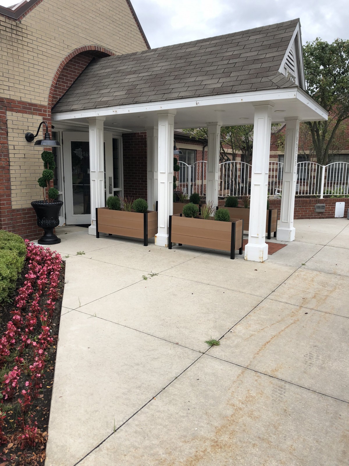 A building entrance with white columns and planters, surrounded by a flower-lined sidewalk.