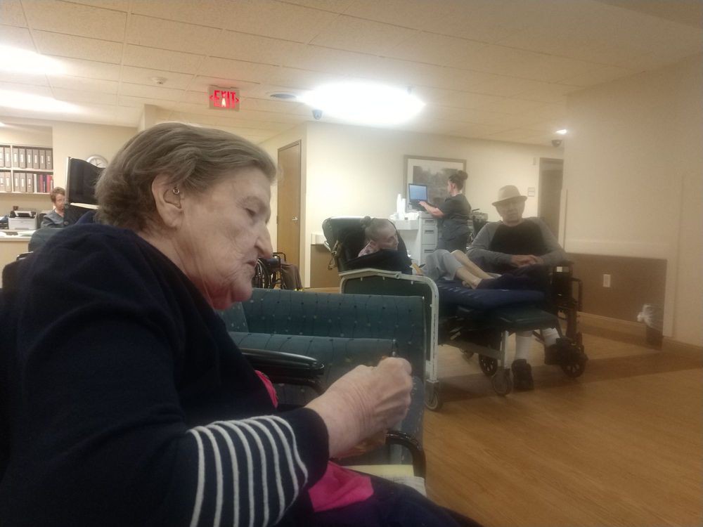 An elderly woman sitting in a nursing home, with others and caregivers in the background.