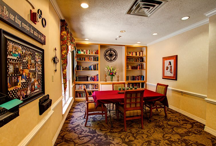 A cozy library room with a red table, surrounded by bookshelves and decorated with artwork.