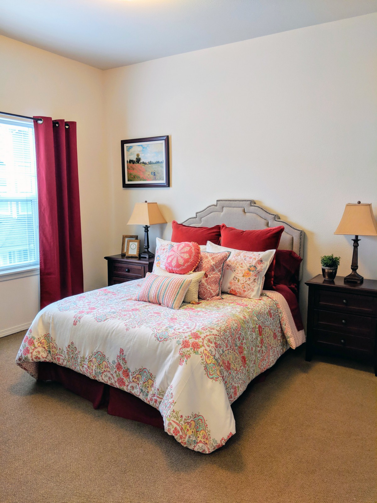 A cozy bedroom with a neatly made bed, decorative pillows, and bedside lamps. The room features a window with red curtains and a framed picture on the wall.