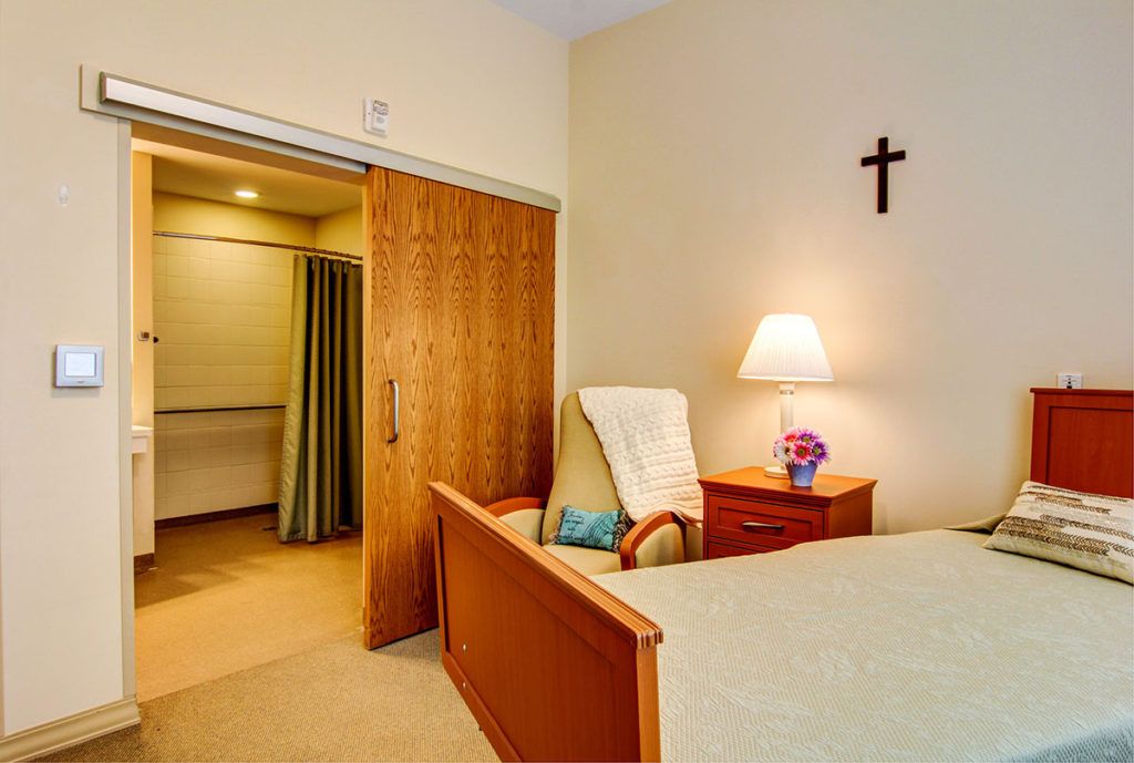 A cozy bedroom featuring a bed, chair, and a lamp, with a cross on the wall.