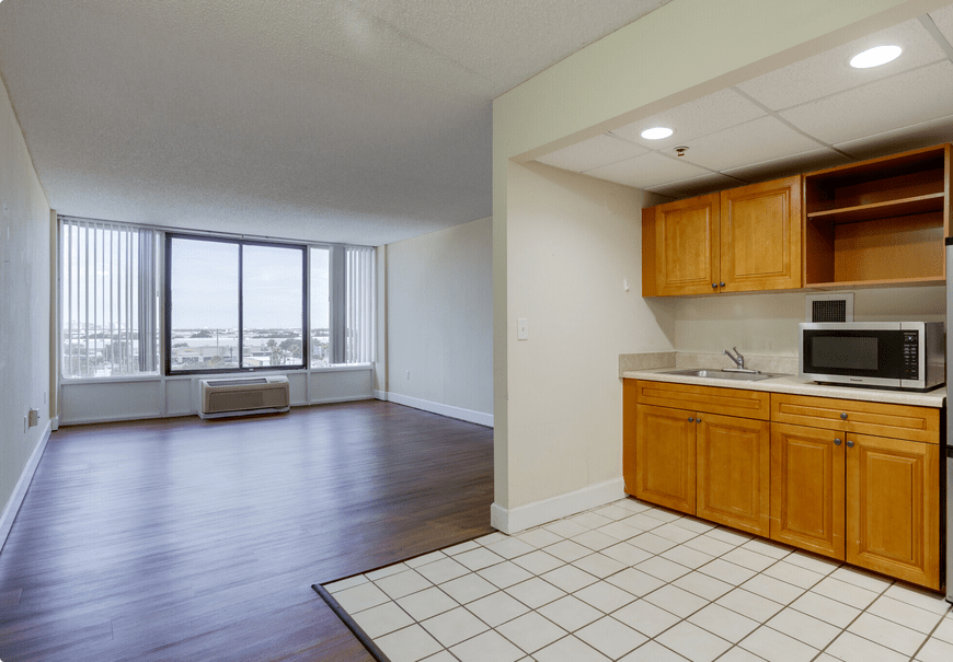 An interior view of an apartment featuring a kitchen with wooden cabinets and a spacious living area with large windows and hardwood flooring.