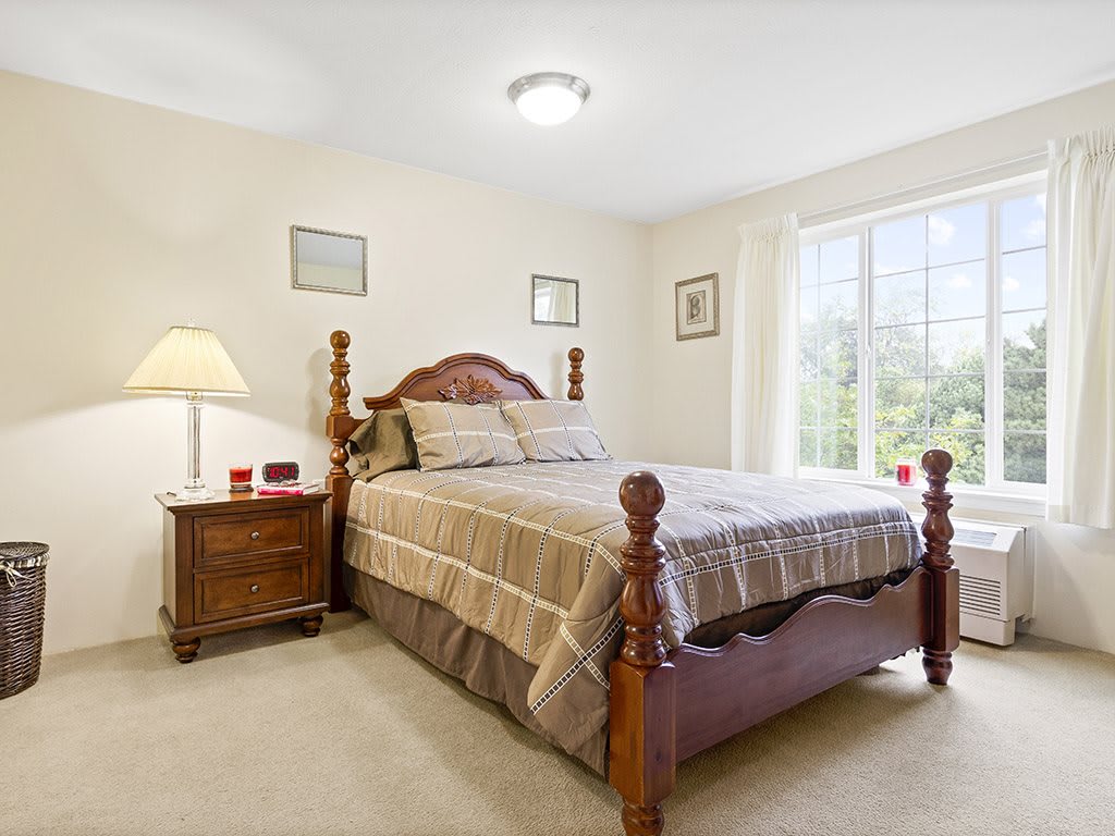 A cozy bedroom with a wooden bed frame, nightstand, and a large window providing natural light.