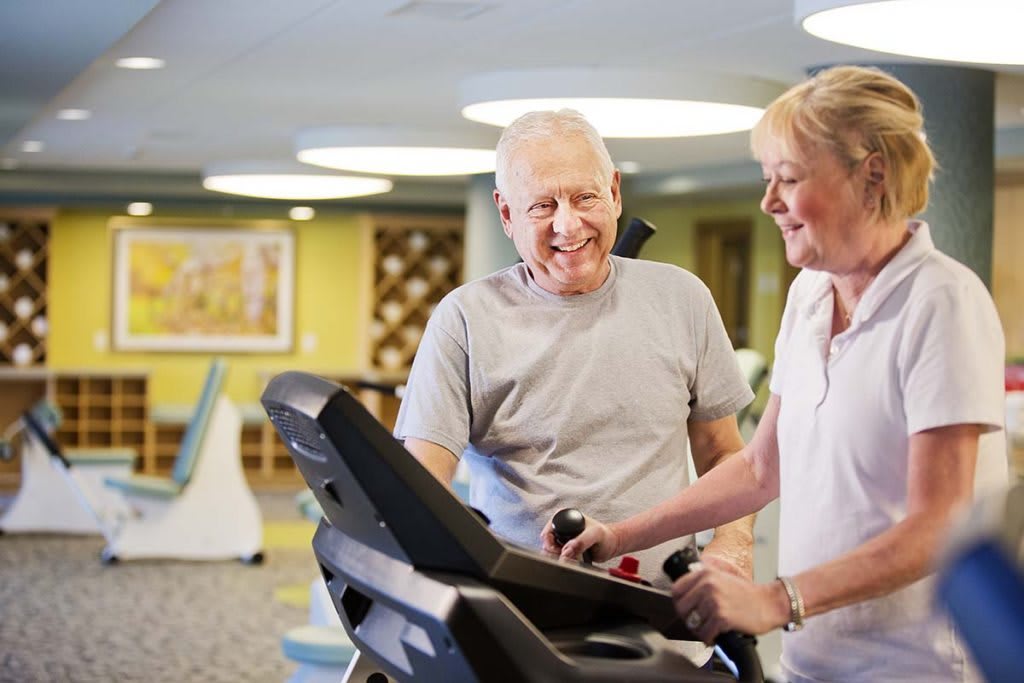 An elderly man receiving assistance while using a treadmill in a gym.