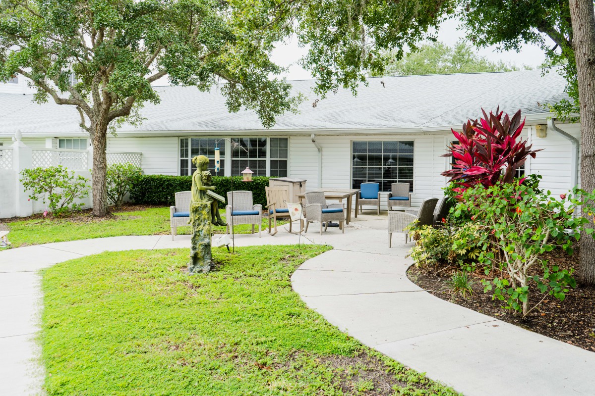 A serene garden setting with a curved walkway leading to outdoor seating in front of a house, surrounded by trees and well-maintained landscaping.