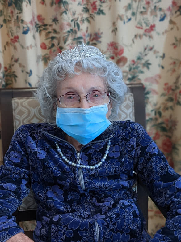 An elderly person sitting indoors wearing a mask and glasses, styled with a pearl necklace and floral-patterned curtains in the background.