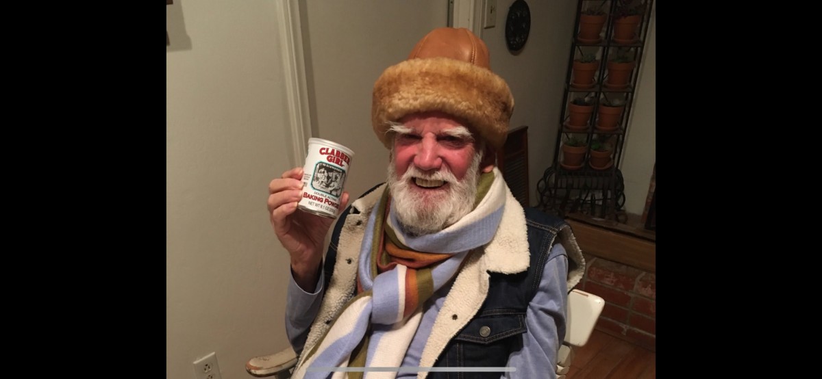 Elderly man dressed warmly, holding a cup indoors.